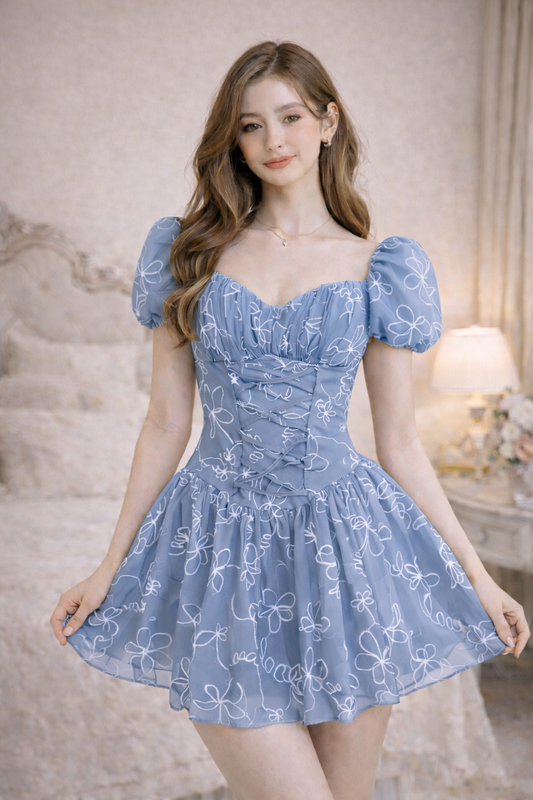 Woman wearing a pastel blue floral embroidered puff sleeve mini dress with lace-up corset front and gathered skirt in a softly lit room