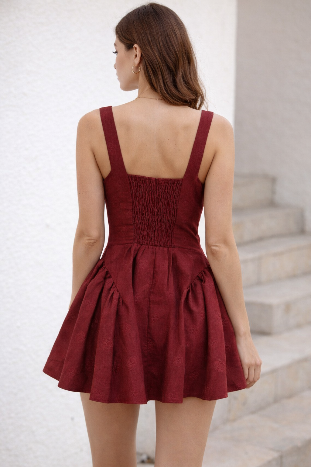 Back view of model in burgundy mini dress displaying elastic shirred panel and flared skirt outdoors